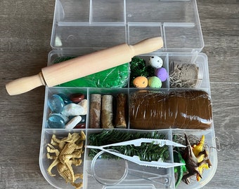 May include: A clear plastic storage box filled with various dinosaur-themed play items, including green and brown play dough, a wooden rolling pin, small plastic dinosaurs, a magnifying glass, tweezers, and small plastic eggs.