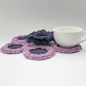 May include: A set of crocheted coasters in shades of purple and gray. The coasters are shaped like flowers and have a center hole for a cup or mug. A white ceramic mug is sitting on top of one of the coasters.