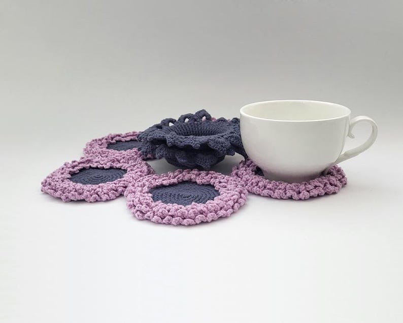 May include: A set of crocheted coasters in shades of purple and gray. The coasters are shaped like flowers and have a center hole for a cup or mug. A white ceramic mug is sitting on top of one of the coasters.