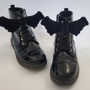 Puede incluir: Un par de botas negras de charol con alas de murciélago de ganchillo negro atadas a los cordones.