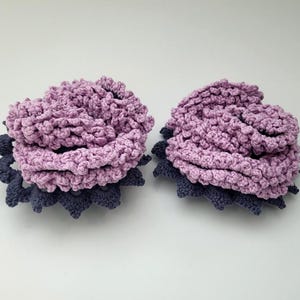 May include: Two crocheted flower hair clips in shades of purple and blue. The flowers are layered and have a textured, ruffled appearance.