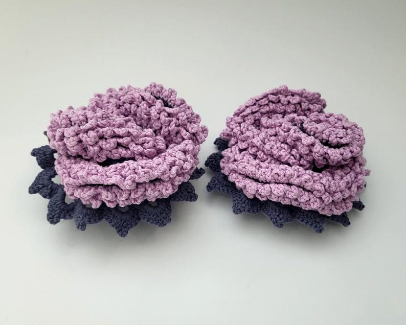 May include: Two crocheted flower hair clips in shades of purple and blue. The flowers are layered and have a textured, ruffled appearance.
