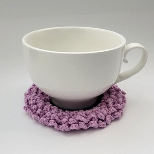 May include: A white ceramic mug with a handle sits on a lavender crocheted coaster.