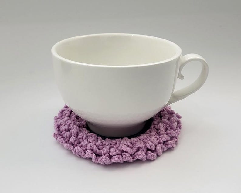 May include: A white ceramic mug with a handle sits on a lavender crocheted coaster.