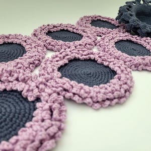 May include: A set of six crocheted coasters with a pink and gray color scheme. Each coaster features a circular design with a raised edge.
