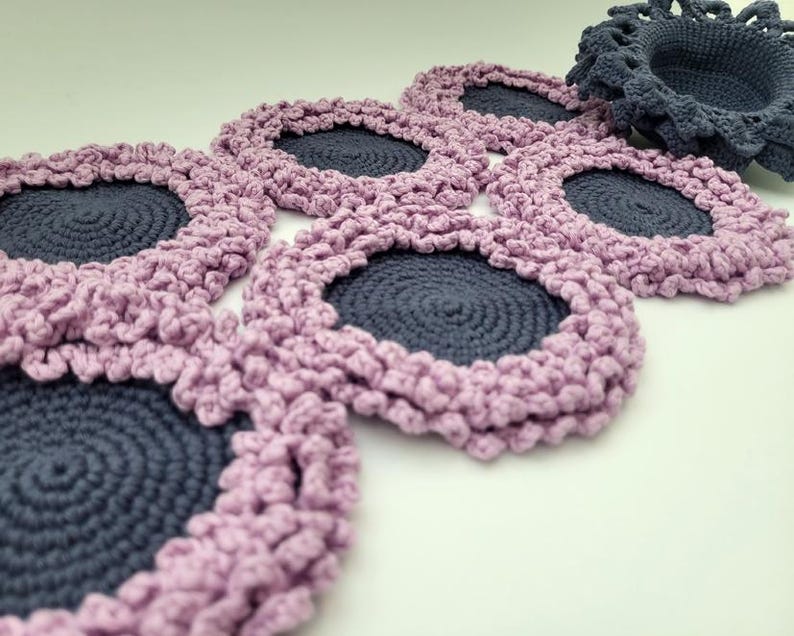 May include: A set of six crocheted coasters with a pink and gray color scheme. Each coaster features a circular design with a raised edge.