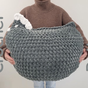 Peut inclure: Un pouf gris en crochet avec un dessus en crochet blanc. Le pouf est tenu par une personne portant un pull marron.