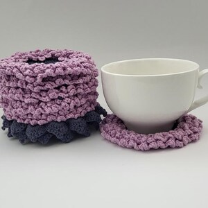 May include: A set of two crocheted lavender and gray coasters with a white ceramic teacup on top of one of the coasters.