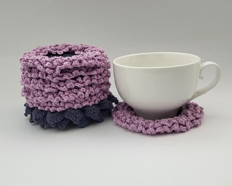 May include: A set of two crocheted lavender and gray coasters with a white ceramic teacup on top of one of the coasters.