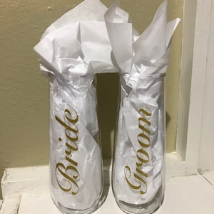 May include: Two clear glass champagne flutes with gold lettering. One flute says "Bride" and the other says "Groom".