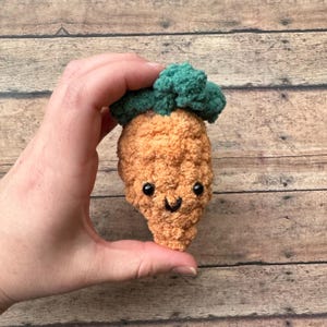May include: A handmade, plush carrot with a smiling face and green top. The carrot is orange with black eyes and a black stitched mouth. The carrot is held in a hand against a wooden background.