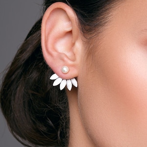 Op de afbeelding: Een vrouw met lang donker haar en blauwe ogen draagt ​​een paar parel- en witte bloemoorbellen. De oorbellen zijn een enkele parel met een cluster van witte bloemblaadjes die naar beneden uitstrekken.