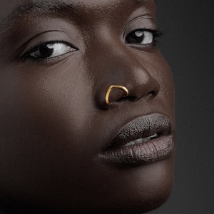 May include: A close-up portrait of a person with short, dark hair wearing a gold nose ring and gold earrings. The person has dark skin and is looking directly at the camera.