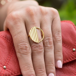 Könnte beinhalten: Ein goldener Ring mit einem Halbkreisdesign mit vertikalen Linien. Der Ring befindet sich an einem Finger einer Hand, die auf einer roten gesteppten Handtasche liegt.