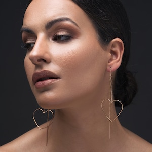 Può includere: Una donna con capelli scuri e trucco indossa lunghi orecchini a forma di cuore dorati. Gli orecchini sono pendenti e hanno una sottile catena.