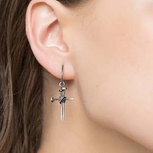 Könnte beinhalten: Silberne Ohrhänger mit einem Kreuzdesign und mehreren Nägeln. Die Nägel sind miteinander verdreht, um die Kreuzform zu bilden.