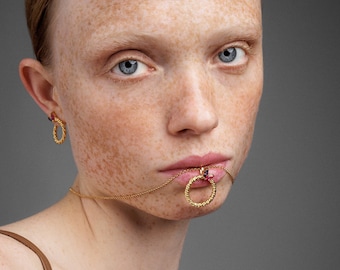 May include: A gold-toned jewelry set featuring a necklace and earrings. The necklace has a circular pendant with a red gemstone, and the earrings have a similar design. The jewelry is worn on a person with fair skin and freckles, against a gray background.