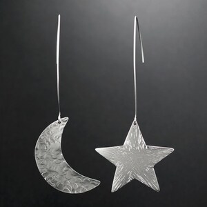 May include: A pair of silver earrings featuring a crescent moon and a star design. The earrings are crafted from hammered metal and have a textured finish.