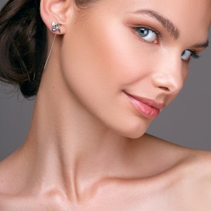 May include: A silver earring with a geometric design is worn in the ear. The earring has a long, thin post that dangles down. The person in the image has fair skin and blue eyes. The background is a solid grey.