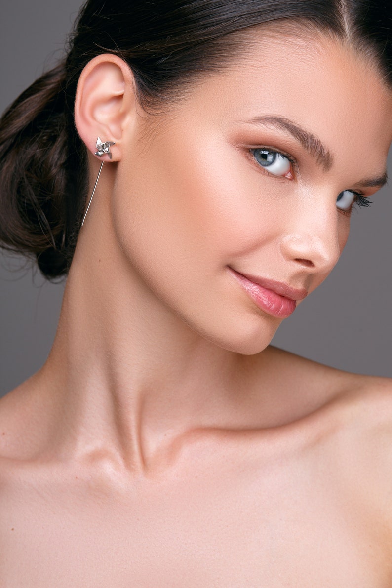 May include: A silver earring with a geometric design is worn in the ear. The earring has a long, thin post that dangles down. The person in the image has fair skin and blue eyes. The background is a solid grey.