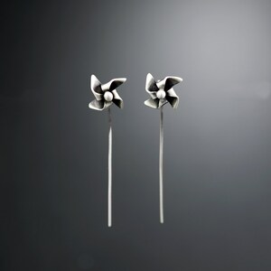 May include: A pair of silver earrings. Each earring has a pinwheel design with a small, round bead in the centre. The pinwheel is attached to a long, slender post. The background is a gradient of grey.