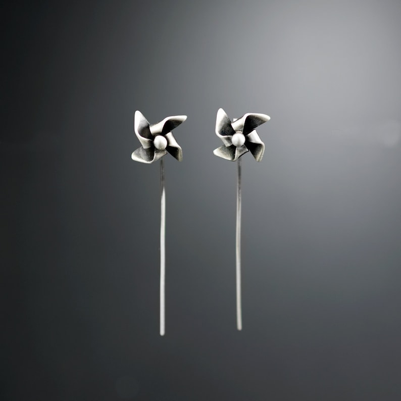 May include: A pair of silver earrings. Each earring has a pinwheel design with a small, round bead in the centre. The pinwheel is attached to a long, slender post. The background is a gradient of grey.