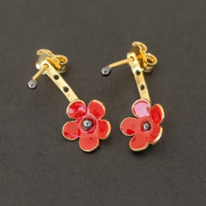 Könnte beinhalten: Ein Paar goldene Ohrringe mit roten Blumenakzenten. Die Ohrringe haben einen langen, dünnen Stiel mit einer kleinen schwarzen Perle oben.