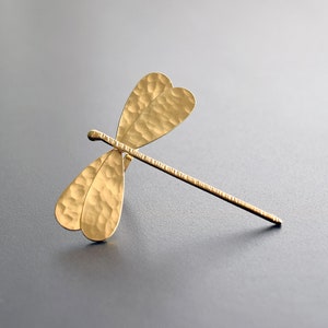 Puede incluir: Un broche de libélula de color dorado con alas texturizadas. La libélula está hecha de metal y tiene un acabado martillado.