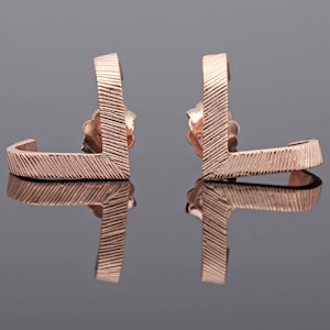 May include: A pair of rose gold earrings with a geometric design. The earrings are made of a textured metal and have a curved shape.
