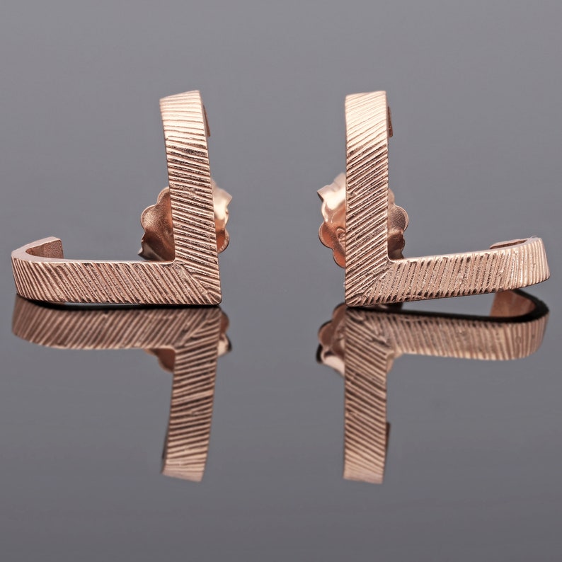 May include: A pair of rose gold earrings with a geometric design. The earrings are made of a textured metal and have a curved shape.