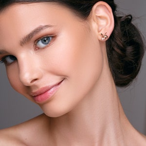 May include: A close-up of a person wearing a gold earring with a geometric design and a long, dangling bar. The earring is on the right ear. The person has fair skin, blue eyes, and dark hair pulled back.