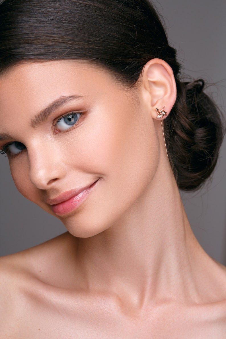 May include: A close-up of a person wearing a gold earring with a geometric design and a long, dangling bar. The earring is on the right ear. The person has fair skin, blue eyes, and dark hair pulled back.