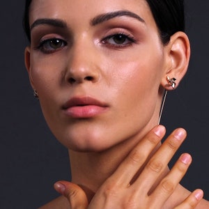 May include: Close-up of a person wearing two earrings. One earring is a small stud, and the other is a gold-coloured, long, dangling earring. The earrings have a floral design with black and gold accents. The background is a solid dark grey.