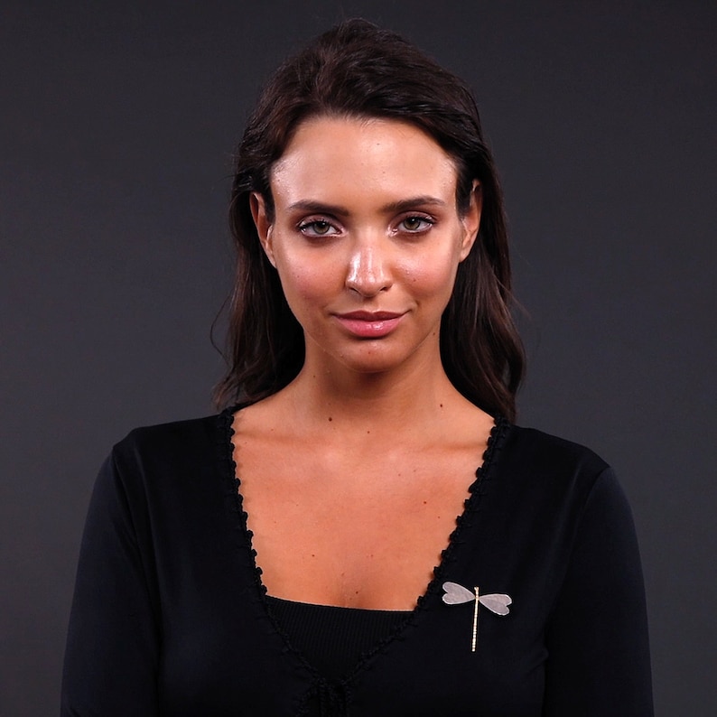 Pode incluir: Uma mulher com cabelo comprido e escuro vestindo um casaco preto com um broche de lib&eacute;lula prateado.
