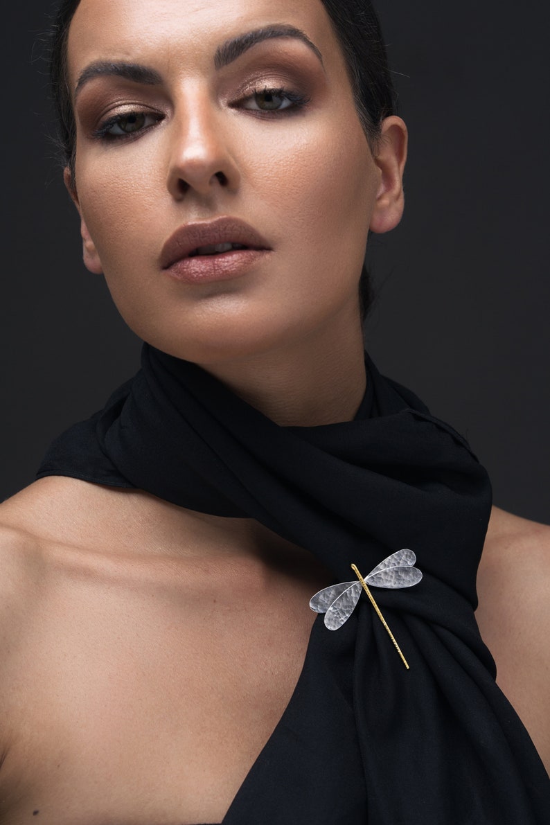 Pode incluir: Uma mulher usando um len&ccedil;o preto com um broche de lib&eacute;lula prateado. A lib&eacute;lula tem um corpo dourado e asas transparentes.