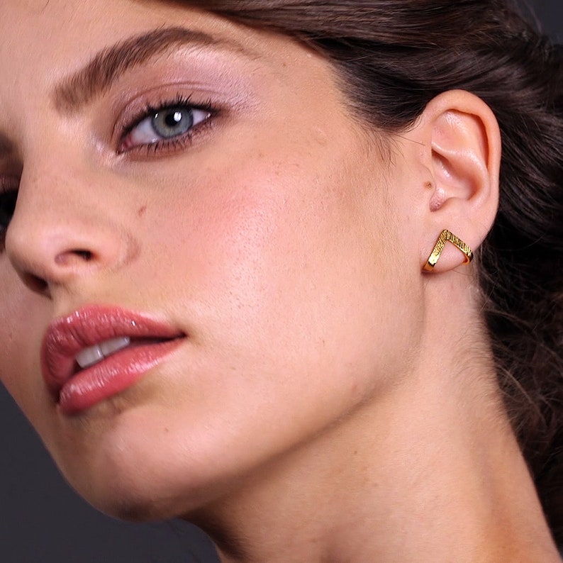 May include: A pair of gold-toned earrings with a V-shaped design. The earrings are made of a textured metal and have a minimalist style.