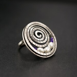 Puede incluir: Un anillo de plata con un diseño en espiral y una fila de perlas blancas y piedras preciosas púrpuras.
