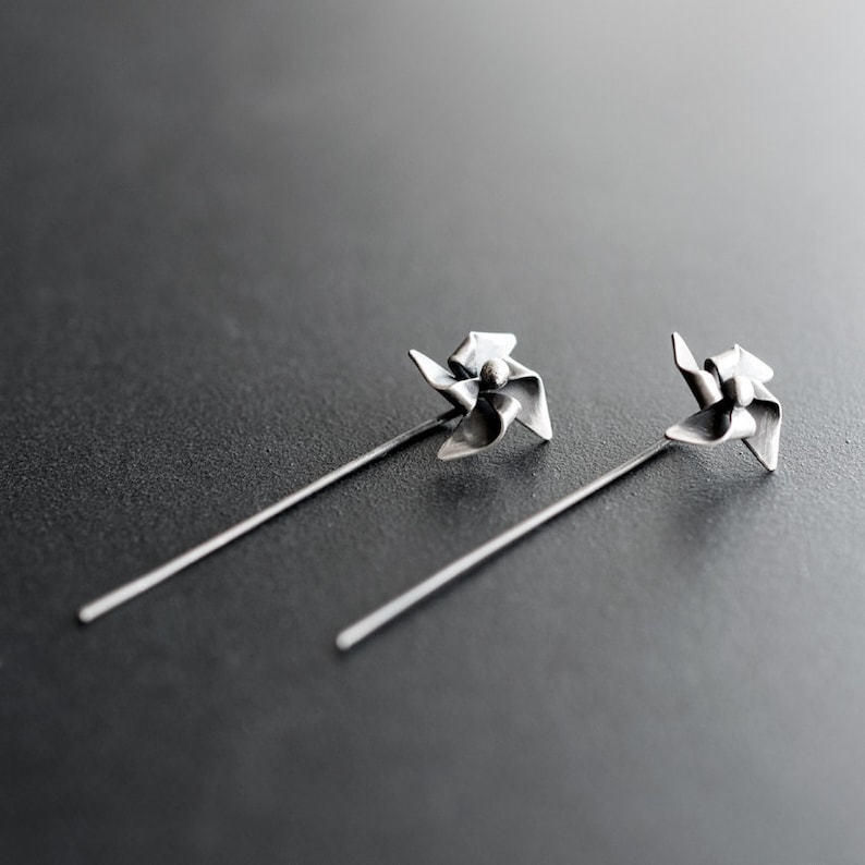 May include: A pair of silver earrings with a pinwheel design. Each earring has a small, detailed pinwheel at the top, attached to a long, slender post. The earrings are set against a dark grey background.