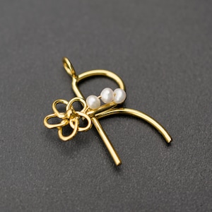 Puede incluir: Collar con colgante de oro en forma de letra R adornado con un pequeño dije de flor y tres perlas blancas.