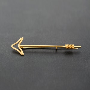 Könnte beinhalten: Eine goldene Pfeilbrosche mit einem gedrehten Drahtdetail am Ende.