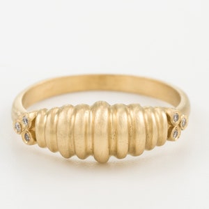 May include: A gold ring featuring a ribbed central design and small diamond accents on either side. The ring has a smooth, matte finish and a classic band. The ring's design is approximately 1.9 cm in length.