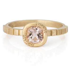 May include: A gold ring featuring a square, pale pink gemstone. The band has a textured design with vertical lines. The gemstone is set in a square bezel with a dotted texture on one side. The ring is shown against a white backdrop.