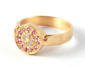 Può includere: Un anello d'oro con un design circolare. Il centro presenta un gruppo di piccole pietre preziose bianche e rosa. L'anello ha una fascia semplice e un'incastonatura decorativa per le pietre preziose. L'anello è su sfondo bianco.