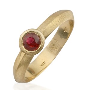 May include: A gold ring with a red ruby gemstone set in a bezel setting. The ring has a textured band and is marked with the initials "WR".