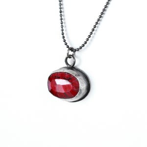 May include: A silver chain necklace with a red gemstone pendant. The pendant is oval shaped and has a silver bezel setting.