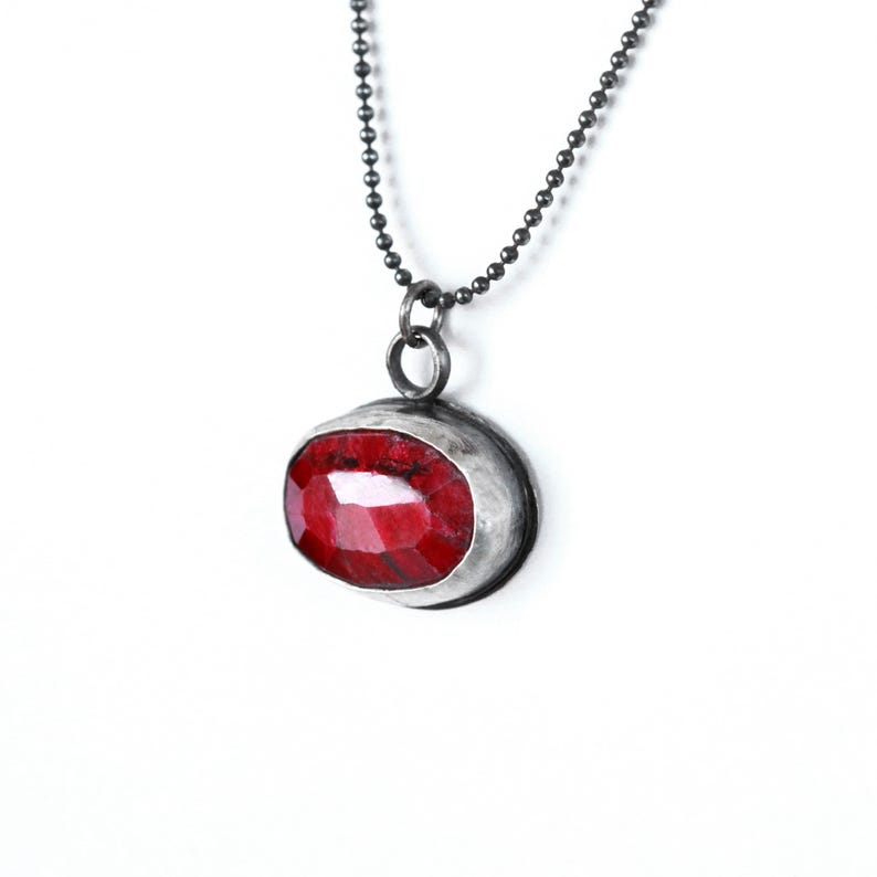 May include: A silver chain necklace with a red gemstone pendant. The pendant is oval shaped and has a silver bezel setting.
