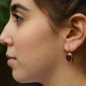 May include: A drop earring featuring a polished oval-shaped deep pink gemstone set in a gold-toned bezel with a dark metallic backing. The earring hangs from a simple silver-toned wire hook, showcasing a smooth, rounded surface and a vibrant color.