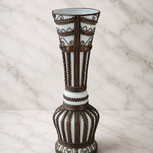 May include: A tall, slender white glass vase featuring an intricate overlay of dark bronze-colored metal filigree. The metalwork forms vertical ribbed patterns, horizontal bands, and decorative scrollwork around the flared rim and the base.