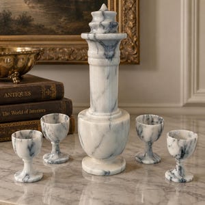 May include: A white and grey marble decanter with a tall, tiered stopper and four matching marble goblets. Each piece features natural veining patterns throughout the stone, with the decanter standing centrally among the four small stemmed cups.