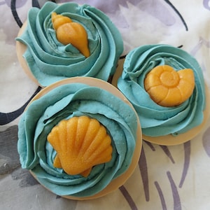 May include: Three round cookies decorated with blue frosting swirls and edible orange seashell shapes. The cookies have a light brown base and are arranged on a patterned surface. The frosting is a vibrant teal color.